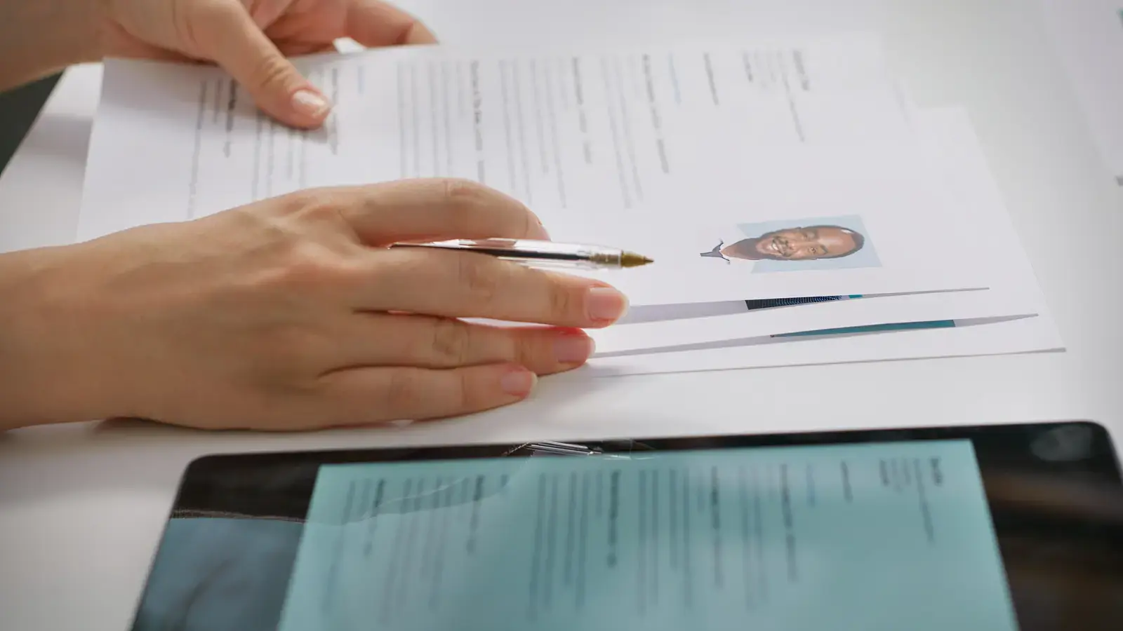 Hands holding a pen over printed CVs on a desk beside a tablet
